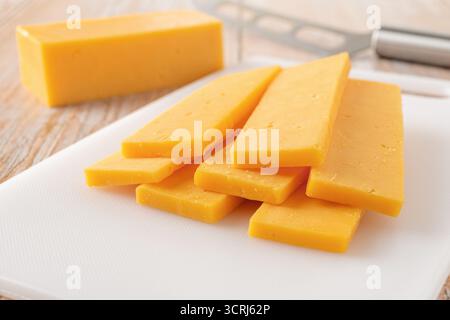 Sliced cheddar cheese close-up. Slaced orange cheese on a white cutting board and knife over rustic wooden tabletop. Dairy product, calcium concept. Stock Photo