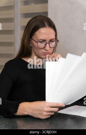 Focused businesswoman is carefully examining documents, possibly reviewing reports, analyzing data, or studying contracts in a home office setting, de Stock Photo