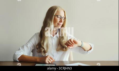Confident mature woman professional working on digital tablet computer with paper documents looks at watch, checks the time, sitting at desk in office Stock Photo