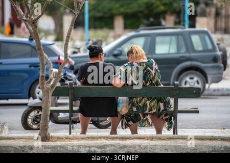 two overweight females sitting on a park bench together viewed from behind. Two large ladies resting on a wooden bench at the roadside. Stock Photo