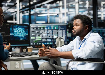 Server hub manager and man in wheelchair use artificial intelligence to perform computing tasks. Engineer with paraplegia and coworker in inclusive data center use AI for machine learning workloads Stock Photo