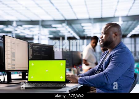 Technician in data center updates rigs systems on green screen laptop to protect servers from exploitable software vulnerabilities. IT professional using chroma key notebook to look for errors Stock Photo