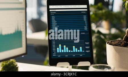 Modern workspace with analytical data paperwork documents in bright office environment with potted plants. Tablet on desk with digital statistics charts displayed on screen, close up shot Stock Photo