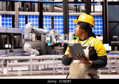 Female industrial engineer walking through power plant with tablet in hand, overseeing machinery development. African American maintenance manager monitors production lines in manufacturing facility. Stock Photo