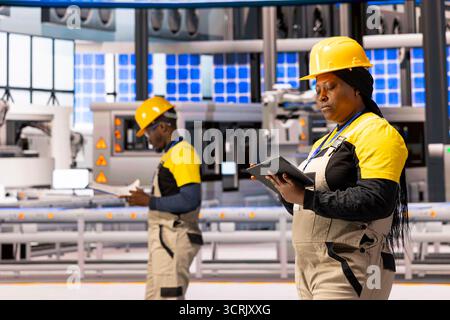 African american technicians inspecting Industry 4.0 plant and assessing manufacturing process. Female engineer holds tablet, conducting smart factory evaluation and quality control. Stock Photo