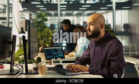 Teamworking colleagues analyzing data charts on computer screen at desk. Team of employees completing spreadsheets on desktop computer, inputting and analyzing data, camera B Stock Photo