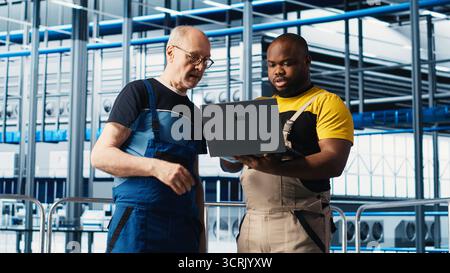 Senior engineer in smart photovoltaics factory talks with intern, showing system performance metrics. Experienced solar panels plant expert guiding new trainee in tracking energy production, camera A Stock Photo