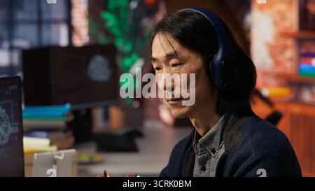 Senior programmer in AI startup office putting on headphones, refining machine learning models to enhance automation. Asian CTO manager listening music while working, camera B close up Stock Photo