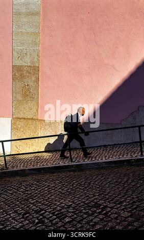 Man strides briskly across sunlit cobblestones, pink wall shadow, urban street scene, Lisbon Stock Photo