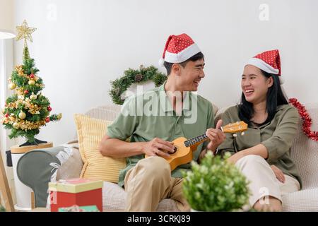 Christmas love, happy couple wears festive hats enjoying winter holiday at home, man plays ukulele Stock Photo