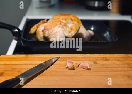 A chicken is being cooked in a pan on a stove. A knife and garlic on a cutting board next to the pan Stock Photo