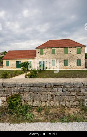 A stone two-story house with green wooden shutters sits in a quiet rural yard. Red-tiled roof, gravel path, and small shrubs create a calm, picturesqu Stock Photo