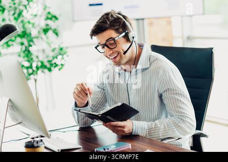 Business professional with headset working productively at desk in office while managing tasks with a notebook and computer Stock Photo