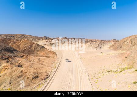 High-angle view of pale gravel road cutting through vast Moon Valley, lone white 4x4 dwarfed by eroded ochre hills and hazy sky, Swakopmund, Namibia. Stock Photo