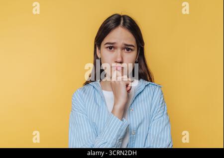 Young woman with uncertain expression poses against bright yellow background while contemplating decisions Stock Photo