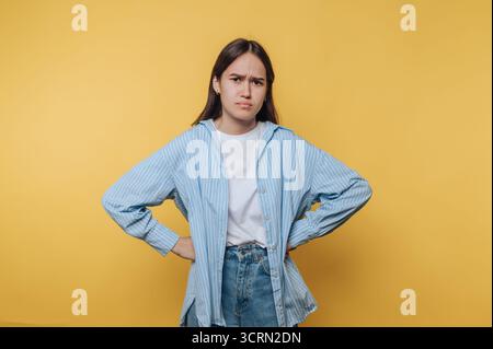 Young woman with a confused expression stands against a bright yellow background while wearing casual attire Stock Photo