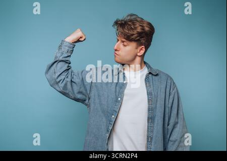 Young man showing off strength and confidence against a solid blue background Stock Photo