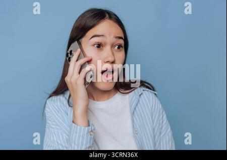 A young woman with long hair engages in a phone conversation, showing surprise on her face. She wears a light blue striped shirt and stands in front o Stock Photo