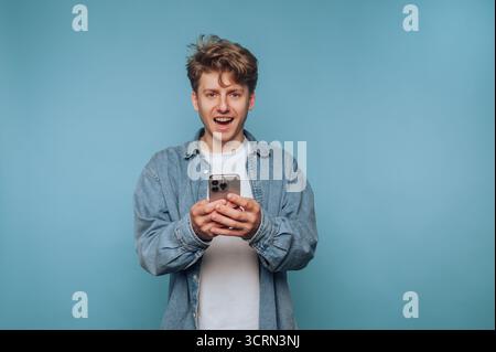 A young man wearing a denim jacket and white shirt smiles while holding a smartphone. He appears cheerful and engaged, standing in front of a blue bac Stock Photo