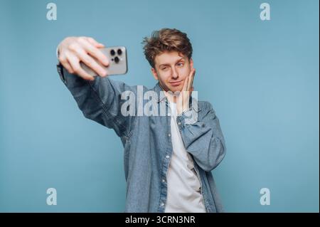 A young man stands confidently in front of a light blue background, taking a selfie. He wears a denim jacket over a white shirt, striking a playful po Stock Photo