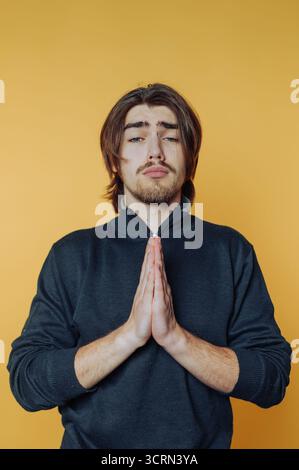A young man stands with hands clasped in front of him, showcasing a look of gratitude. The bright yellow background enhances his expression of appreci Stock Photo