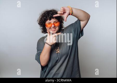 A person with curly hair wearing orange sunglasses and a gray t-shirt poses playfully, framing their face with their hands against a plain background. Stock Photo