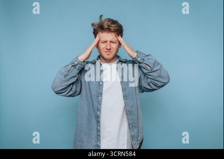 A young man in a denim shirt and white t-shirt stands against a blue background, holding his head with both hands and expressing discomfort or stress. Stock Photo