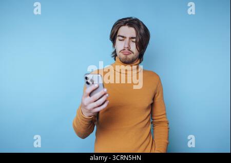 Young man in orange sweater looking at smartphone with puzzled expression. Stock Photo