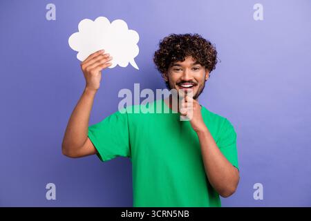 Young man in green shirt against purple background holding cut-out speech bubble, expressing thoughtfulness and creativity Stock Photo