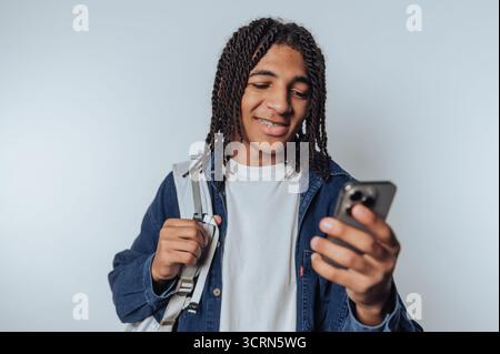 Young man with braided hair smiling while looking at smartphone, carrying a backpack, wearing a denim jacket and white t-shirt, standing against a pla Stock Photo