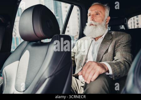 Senior businessman with beard sitting in car's backseat on urban street, dressed elegantly in jacket Stock Photo