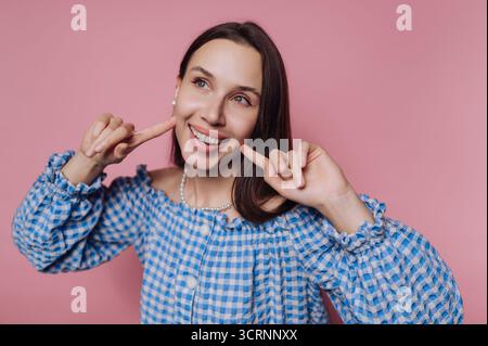 A cheerful woman in a blue checkered blouse poses against a pink background, smiling and pointing at her cheeks. Stock Photo
