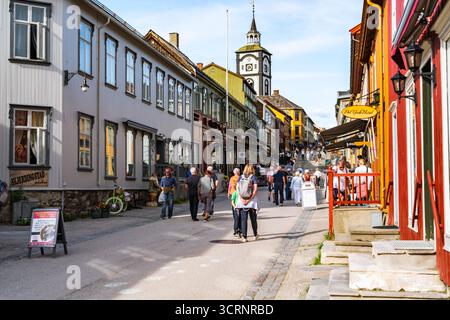 Tourists walk along the charming shopping street of Røros, Norway, taking in the historic buildings and vibrant atmosphere on a pleasant summer day. Stock Photo
