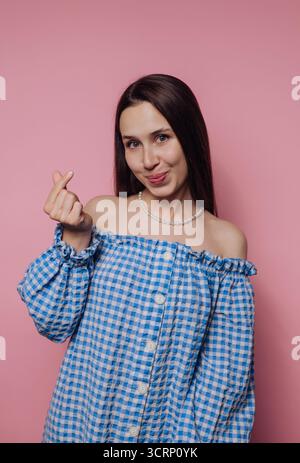 A young woman poses confidently with a playful gesture, wearing a blue checkered off-shoulder top. The vibrant pink background enhances her cheerful e Stock Photo