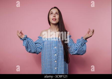 A woman stands gracefully with her hands lifted as if in meditation, adorned in a blue and white gingham dress. She smiles gently while a pink backdro Stock Photo