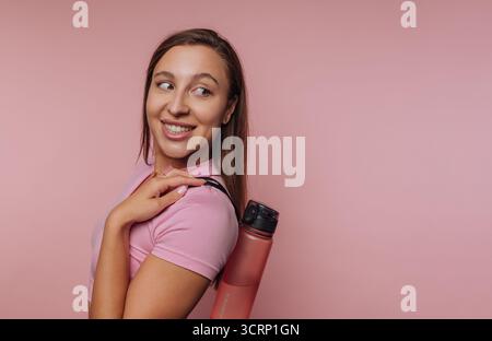 A woman is smiling confidently while holding a water bottle against her shoulder. She is dressed in a pink shirt, standing in front of a vibrant pink Stock Photo