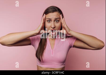 A young woman stands against a soft pink backdrop, her expression one of shock and amazement. She holds her head with both hands, conveying strong emo Stock Photo