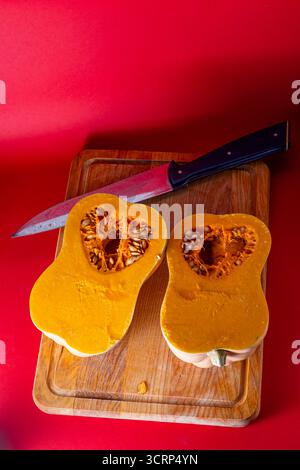 Top view of a fresh butternut squash cut in half on a wooden cutting board with visible seeds and pulp. A large kitchen knife rests nearby against a r Stock Photo