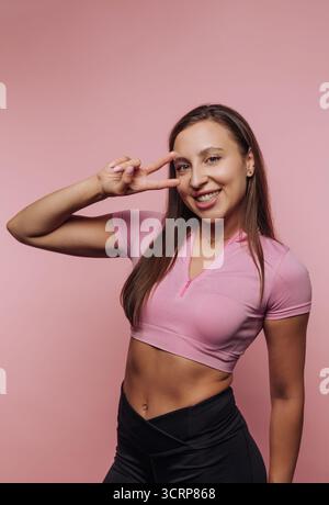 A young woman posing with a peace sign against a pink background, wearing a cropped pink top and black pants, smiling confidently Stock Photo