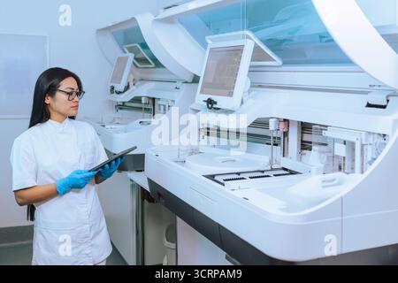 Female technician in a medical laboratory using modern technology in diagnostic process, a tablet and an automated hematology analyzer. Stock Photo