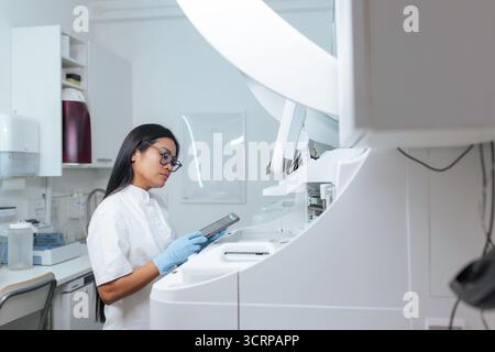 Laboratory technician, young Asian woman holding a tablet and monitoring the workflow and results of an automated digital chemical analyzer. Stock Photo