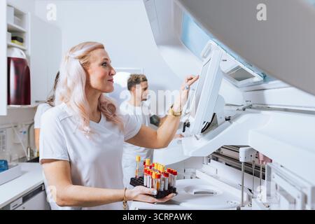 A laboratory technician is organizing blood samples while a colleague works on equipment in a bright lab. Stock Photo
