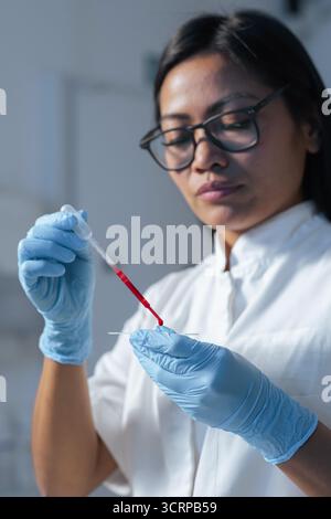 Female laboratory technician pipetting blood sample on the slide, preparing specimen for microscope examination. Blood testing concept. Stock Photo
