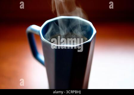 A tall dark ceramic mug filled with hot coffee sits on a wooden windowsill. Steam rises in natural daylight, creating a cozy refreshment scene. Stock Photo