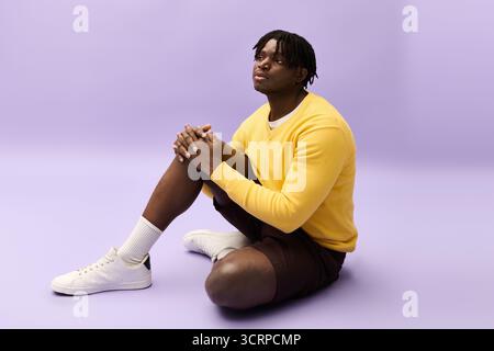 A young man in a yellow sweater and shorts sits thoughtfully on the floor. Stock Photo