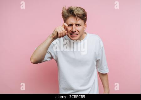 A young man with blond hair shows annoyance by pointing towards the camera. He stands in front of a solid pink background, conveying a strong emotiona Stock Photo