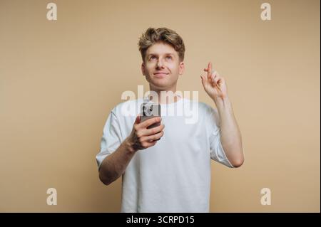 A young man is holding a smartphone in his left hand while pointing upward with his right hand and smiling. He appears to be in a light-colored room, Stock Photo