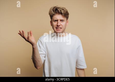 Young man stands against a beige background, looking perplexed with his hand raised, showcasing a mix of confusion and frustration. His casual attire Stock Photo