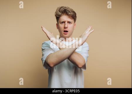A young man stands with a surprised expression, arms crossed in front of his body. He appears to be refusing or rejecting something. The background is Stock Photo