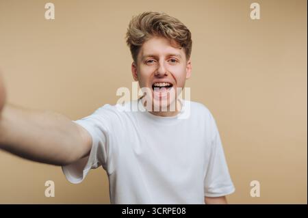 A cheerful young man with light blonde hair is enthusiastically smiling and reaching out with his arm. He wears a plain white t-shirt and appears happ Stock Photo